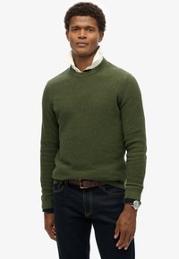 Khakigrüner Pullover mit einer strukturierten, gerippten Musterung, über einem weißen Hemd mit Kragen getragen. Kombiniert mit dunkelblauen Jeans und einem braunen Gürtel.