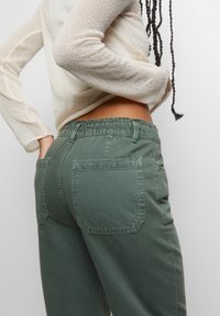 Pantalon vert olive à taille élastique avec deux poches arrière et une coupe décontractée. Le tissu semble doux avec un léger éclat.