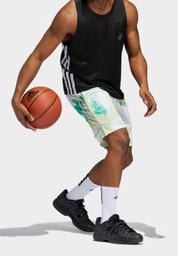 Top de tirantes negro de malla y pantalones cortos vibrantes con un patrón en verde y azul, combinados con zapatillas deportivas negras y calcetines blancos, driblando un balón de baloncesto.