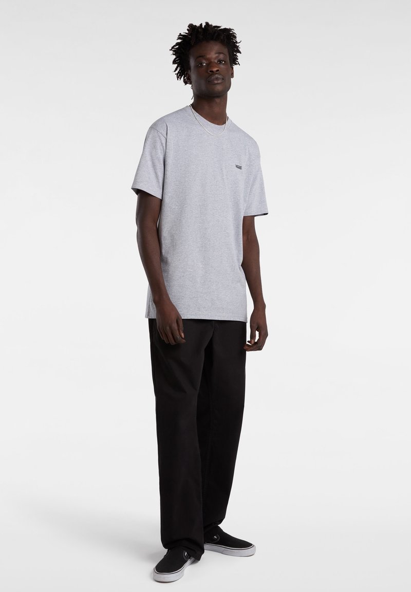 Jeune homme debout portant un t-shirt gris clair à manches courtes, un pantalon noir et des chaussures noires slip-on sur fond blanc.