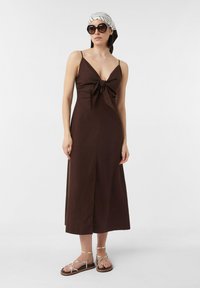 Braunes Baumwoll-Maxi-Kleid mit einem Knoten-Detail vorne, dünnen Trägern und einem V-Ausschnitt. Kombiniert mit sandalen mit goldenen Riemen und einem gemusterten Kopftuch.