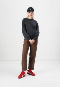 Hanorac negru cu mâneci largi, pantaloni maro și adidași roșii. Îmbrăcămintea are un guler rotund și un aspect casual.