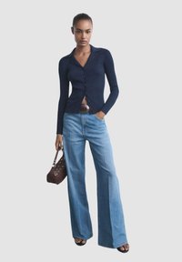 Marineblå ribbet cardigan med krave, knaplukning og flagrende lyseblå jeans. Tilbehør inkluderer en brun håndtaske og et bredt hvidt bælte.