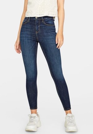 Kvinde iført mørkeblå high-waisted skinny jeans, hvide chunky sneakers og en ærmeløs hvid blondebluse, stående mod en ensfarvet baggrund.