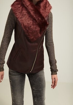 Person wearing dark red faux fur collar vest with zipper over patterned long-sleeve top and faded gray jeans, standing against plain background.