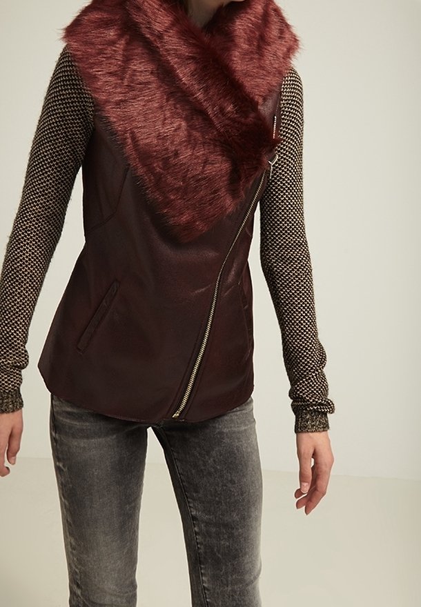 Personne portant un gilet à col en fausse fourrure rouge foncé avec fermeture éclair sur un haut à manches longues à motifs et un jean gris délavé, debout devant un fond uni.