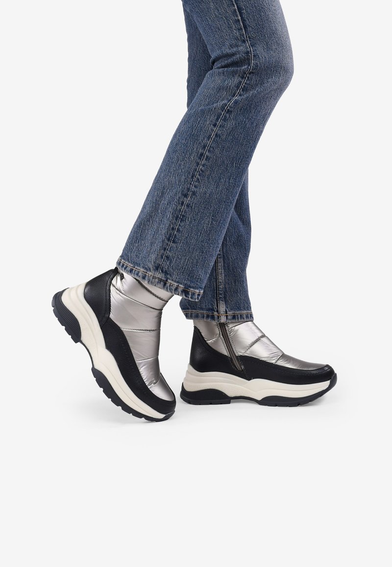 Botines negros y plateados con un acabado brillante, que cuentan con cierre de cremallera y suela blanca gruesa, usados con jeans de mezclilla azules.