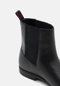 Bottine Chelsea noire en cuir lisse, avec des panneaux élastiques sur les côtés et une languette rouge à l'arrière. Le design présente un bout arrondi et un talon bas.