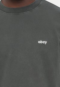 Grafisk t-skjorte i mørkegrå bomull med teksturert overflate, som har en liten hvit "obey" logo på brystet.