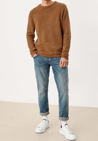 Hnědý pletený svetr s kulatým výstřihem, doplněný o světle modré zúžené džíny. Bílé tenisky s černými akcenty a ohrnuté nohavice dotvářejí celý outfit.