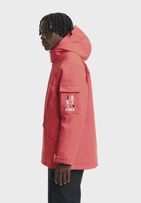 Vyras, stovintis šonu, vilkintis koralinį džemperį su kapuce ir ženkleliu viršutinėje rankovėje, bei juodas kelnes prieš paprastą šviesų foną.