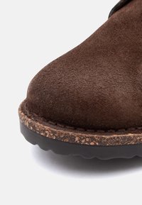 Κοντινό πλάνο της μύτης καφέ δερμάτινου μποτάκι με υφή φελλού στη μεσαία σόλα και μαύρη καουτσούκ εξωτερική σόλα σε λευκό φόντο.