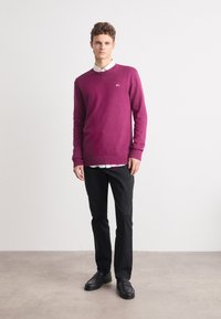 Lilla strikket genser med ribbestrikket mansjetter og hem, brukt over en hvit skjorte med krage. Kombinert med svarte slim-fit bukser og svarte sko.