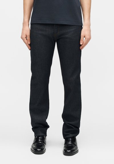 Homme portant un jean bleu foncé, des chaussures habillées en cuir noir et une chemise bleu marine, debout les bras détendus le long du corps sur un fond uni.