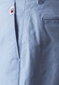 Lyseblå bomullsshorts med glidelås, flat front, sidelommer og et lite merke med røde og blå detaljer.