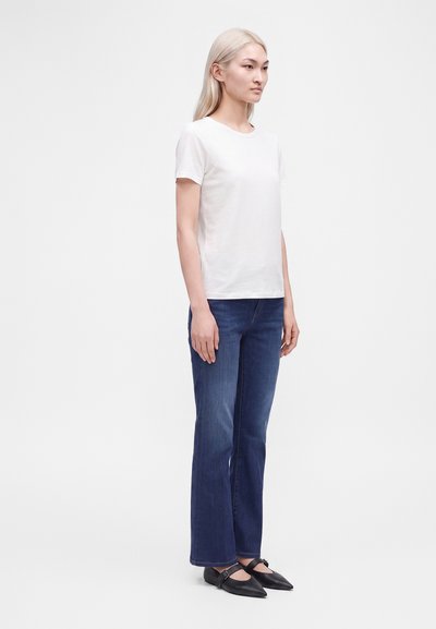 Femme aux longs cheveux blonds portant un t-shirt blanc à manches courtes, un jean bleu droit et des ballerines noires pointues, vue de profil.