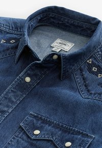 Camisa de mezclilla azul oscuro con cuello, bordado decorativo cerca de los hombros y cierres de presión en la tapeta y el bolsillo.