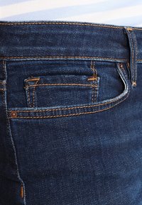 Tmavě modré džíny z denimu s zadní kapsou s oranžovým prošíváním a kovovým nýtem, které vyzdvihují hladkou texturu a přiléhavý střih.