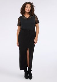Blusa nera con maniche corte in pizzo e scollatura a V, abbinata a una gonna nera a vita alta con spacco frontale, completata da stivaletti neri alla caviglia.