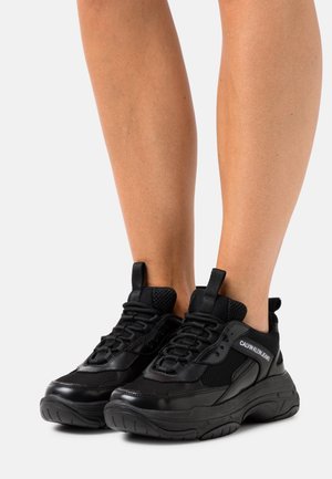Jambes portant des baskets épaisses Calvin Klein Jeans noires avec des détails en mesh et en cuir sur fond blanc.