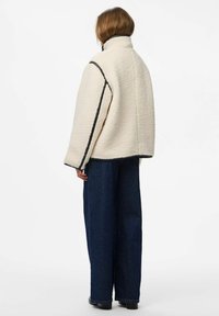 Chaqueta extragrande color crema en tejido texturizado, con acentos negros a lo largo de las costuras. Estilizada con vaqueros oscuros de pierna ancha.