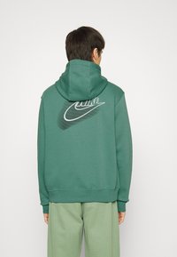 Person som bär en grön Nike-hoodie med en stor vit och svart Nike-logga på baksidan, ihop med ljusgröna byxor, vänd bort.