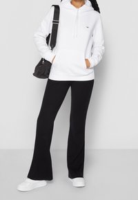 Witte sweatshirt met een capuchon, een voorzak en een klein logo; gecombineerd met zwarte geribbelde uitlopende broeken en witte sneakers met een platte zool.