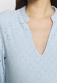 Blusa di cotone azzurro chiaro con scollatura a onda, ricami ad occhio di bue e un motivo geometrico testurizzato. Design elegante e traspirante.