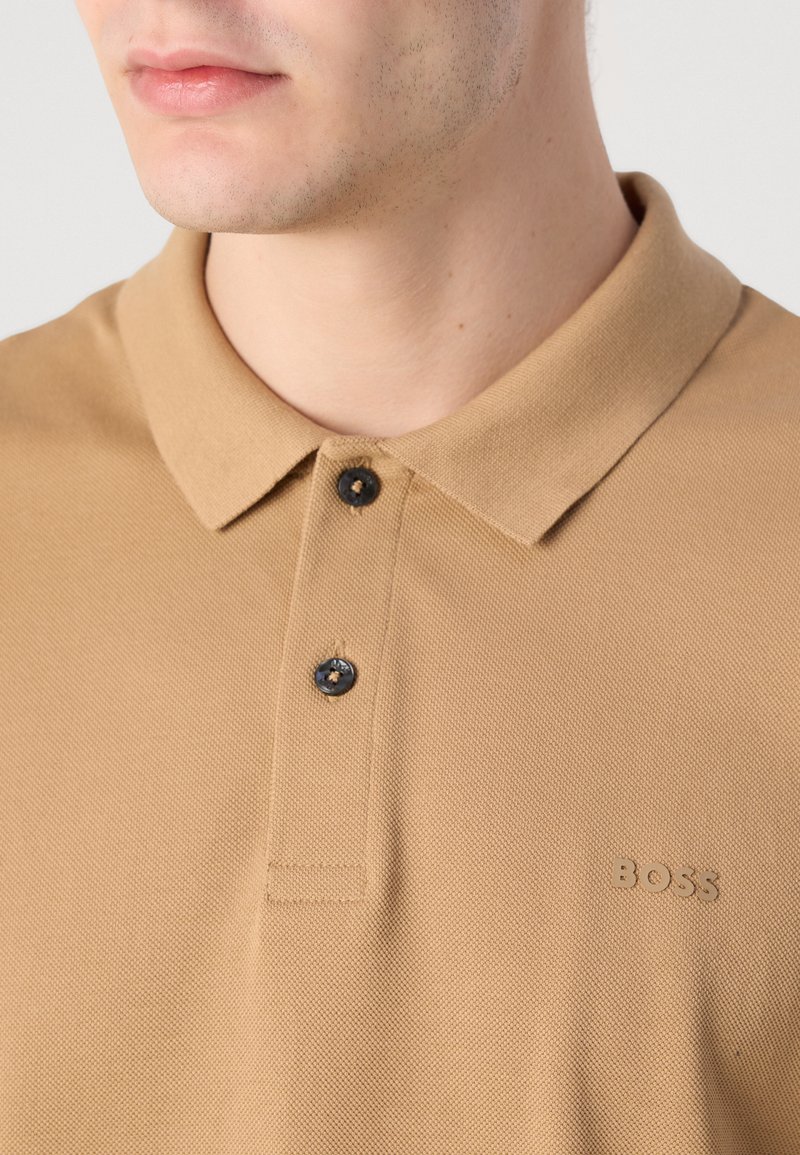 Polo shirt beige en tissu texturé, avec un col, deux boutons noirs et un petit logo "BOSS" en relief sur la poitrine.