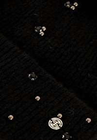 Tessuto di maglia nera con una texture a costine, adornato con accenti argentati e gemme nere di varie dimensioni, inclusi dettagli di logo rotondo.