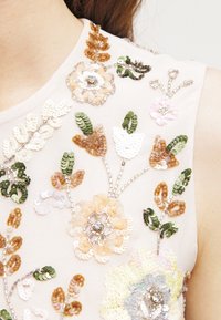 Broderad ärmlös topp i mjukt rosa tyg med blommönster i paljetter och pärlor i färger som grönt, beige och pastellnyanser.