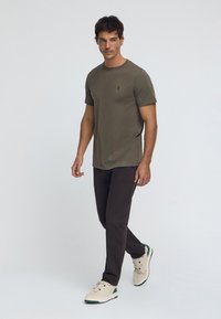 T-shirt manches courtes vert olive avec un col rond, associé à un pantalon marron foncé et des baskets beiges avec des accents verts.