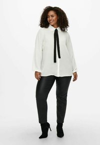 Blusa blanca de manga larga con cuello puntiagudo y corbata negra, combinada con pantalones de cuero negro ajustados y botas de tobillo de tacón.