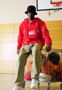 Röd vindjacka med "BULLS"-logotyp, beiga byxor med tryckknappar, vita sneakers, sittande på en trådvagn fylld med basketbollar.