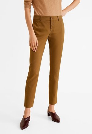 Schmal geschnittene braune Hose mit fünf vorderen Knöpfen, kombiniert mit einem in die Hose gesteckten beigen Pullover und dunkelbraunen spitzen Pumps.