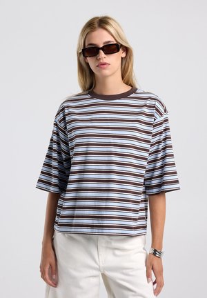 Vrouw draagt rechthoekige donkere zonnebril, een losse blauwbruine gestreepte korte mouwen blouse, witte broek en een zilveren armband.