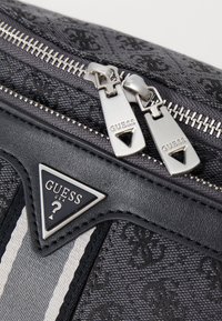 Black fabric bag featuring a textured pattern, silver zippers, and a triangular logo with "GUESS USA." Striped accents in gray and white.
