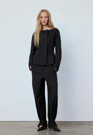 Blonde vrouw draagt een zwarte asymmetrische jas, bijpassende zwarte broek die bij de enkels is samengebonden, en zwarte puntige schoenen tegen een effen achtergrond.