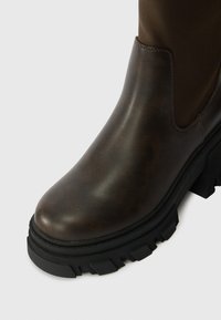 Καφέ δερμάτινη μπότα αστράγαλου με ματ φινίρισμα, χοντρή μαύρη καουτσούκ σόλα και διακριτική γυαλάδα. Περιλαμβάνει στρογγυλή μύτη και πλαϊνό ελαστικό πάνελ.