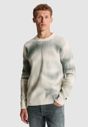 Jeune homme aux cheveux bouclés portant un pull tie-dye blanc et gris à manches longues et un jean bleu, debout avec une main dans sa poche.