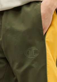 Χέρι ατόμου στην τσέπη παντελονιού ελιάς και κίτρινο με το λογότυπο Champion κοντά στην άκρη της τσέπης.