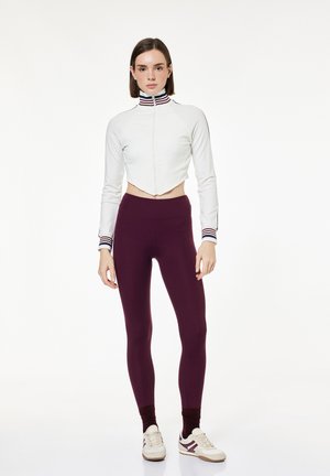 Jonge vrouw die een witte cropped jas met ritssluiting, gestreepte kraag en manchetten draagt, donkerpaarse leggings en beige sneakers, staat stil.