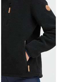 Veste en polaire noire avec une surface texturée et duveteuse. Comprend une poche zippée et un patch en cuir sur la manche gauche.