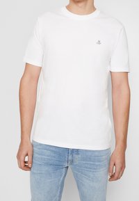 T-shirt bianca in cotone con scollatura tonda, maniche corte e un piccolo logo nero sul petto sinistro. Abbinata a jeans di denim blu chiaro.
