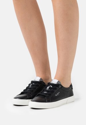 Mustat nahkaiset tennarit, joissa on valkoiset kumipohjat, mustat nauhat ja hopeiset yksityiskohdat kielekkeessä ja korossa. Sileä pinta ja minimaalinen brändäys.