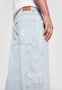 Světle modré denimové cargo kalhoty mají vyšitý logo, hluboké kapsy a koženou nášivku na pásku. Viditelné detaily texturované tkaniny.