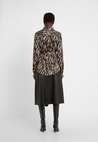 Femme portant un chemisier beige et noir à imprimé animal, rentré dans une jupe midi en cuir noir, avec des bottes à talons noires montant jusqu'aux genoux.