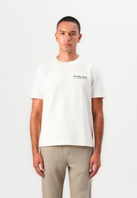 T-shirt en coton crème à manches courtes avec un logo vert foncé sur la poitrine gauche. Col rond simple et coupe décontractée.