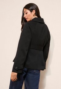 Musta teksturoidusta kankaasta valmistettu takki, jossa on jämäkkä kaulus, vyötäröllä vyö ja napilliset hihansuut. Päällä vaaleanvärisen topin päällä.