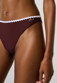 Vínové plavkové nohavičky so bielym zvlneným lemom a výšivkou Karl. Hladká textúra, nízky pás zdôrazňujúci boky.